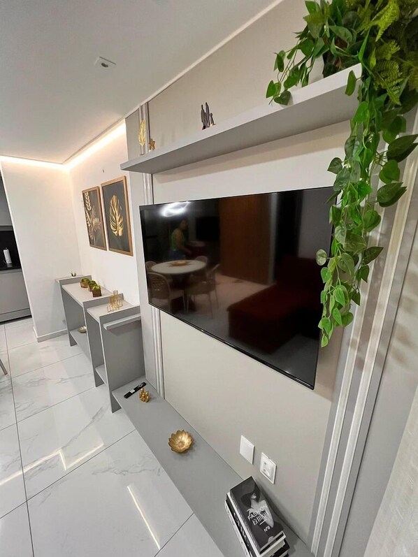 Una sala moderna con un televisor de pantalla plana grande montado en la pared, una mesita redonda con sillas, una planta decorativa y algunas obras de arte enmarcadas en la pared.