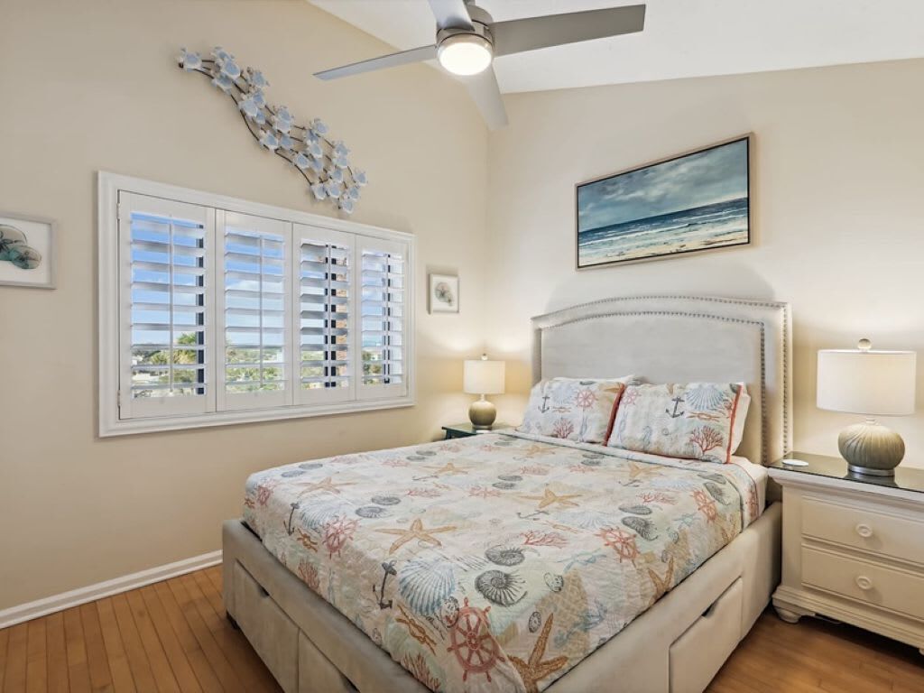 A bedroom with a bed, a ceiling fan, a window with blinds, a nightstand, and a painting on the wall.