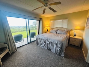 A bedroom with a bed, a ceiling fan, a nightstand with a lamp, a chair, and a view of the outdoors.
