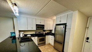 A kitchen with white cabinets, a black countertop, stainless steel appliances, and beige tiled flooring. - Oceanview 2 BR Unit 411 (St. Augustine)