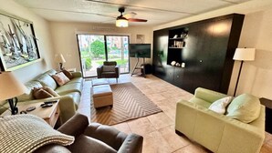 A living room with a sofa, armchair, and a television. There is a bookshelf and a window with a view of the outdoors. - Oceanview 2 BR Unit 411 (St. Augustine)