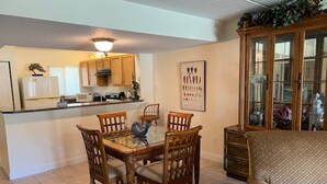 A dining area with a glass-top table and wooden chairs, a kitchen with wooden cabinets and a white refrigerator, and a patterned sofa in the living area.