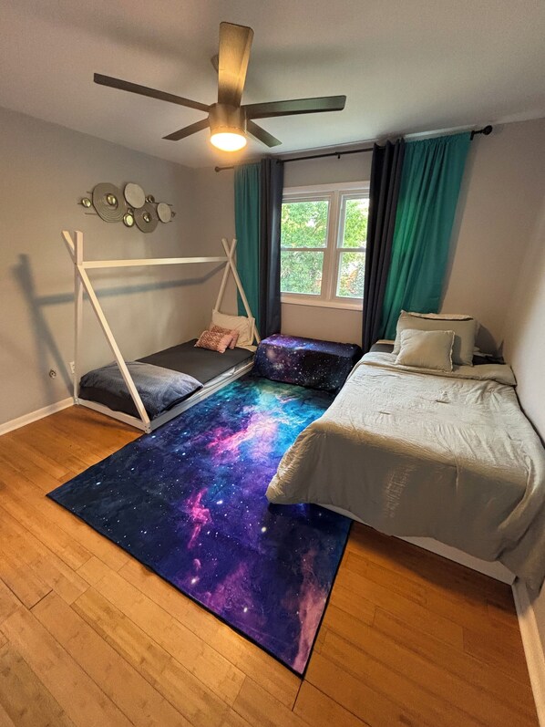 A bedroom with a galaxy-themed rug, a bed, a daybed, a ceiling fan, and a window with curtains.