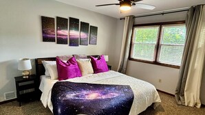 A bedroom with a bed, a ceiling fan, a nightstand, a window with curtains, and wall art. - Galactic Oasis Wonderful 5 bedroom theme home, Wi-Fi, king beds and smart tvs (Aurora)