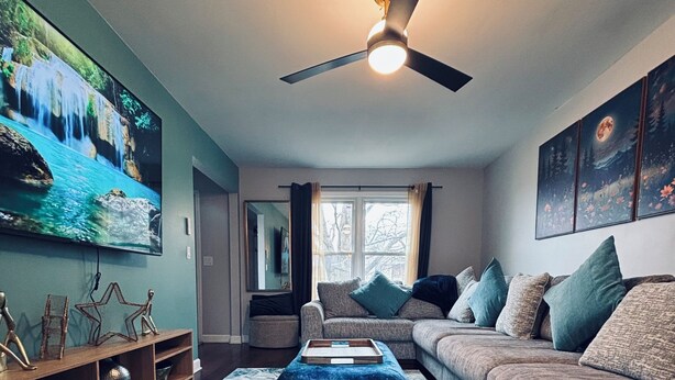 A modern living room with a large flat-screen TV, a ceiling fan, a sofa with multiple cushions, and a patterned rug. - Galactic Oasis Wonderful 5 bedroom theme home, Wi-Fi, king beds and smart tvs (Aurora)