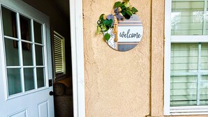 Pared exterior color beige con un letrero de 'welcome', una puerta blanca y una ventana con persianas.