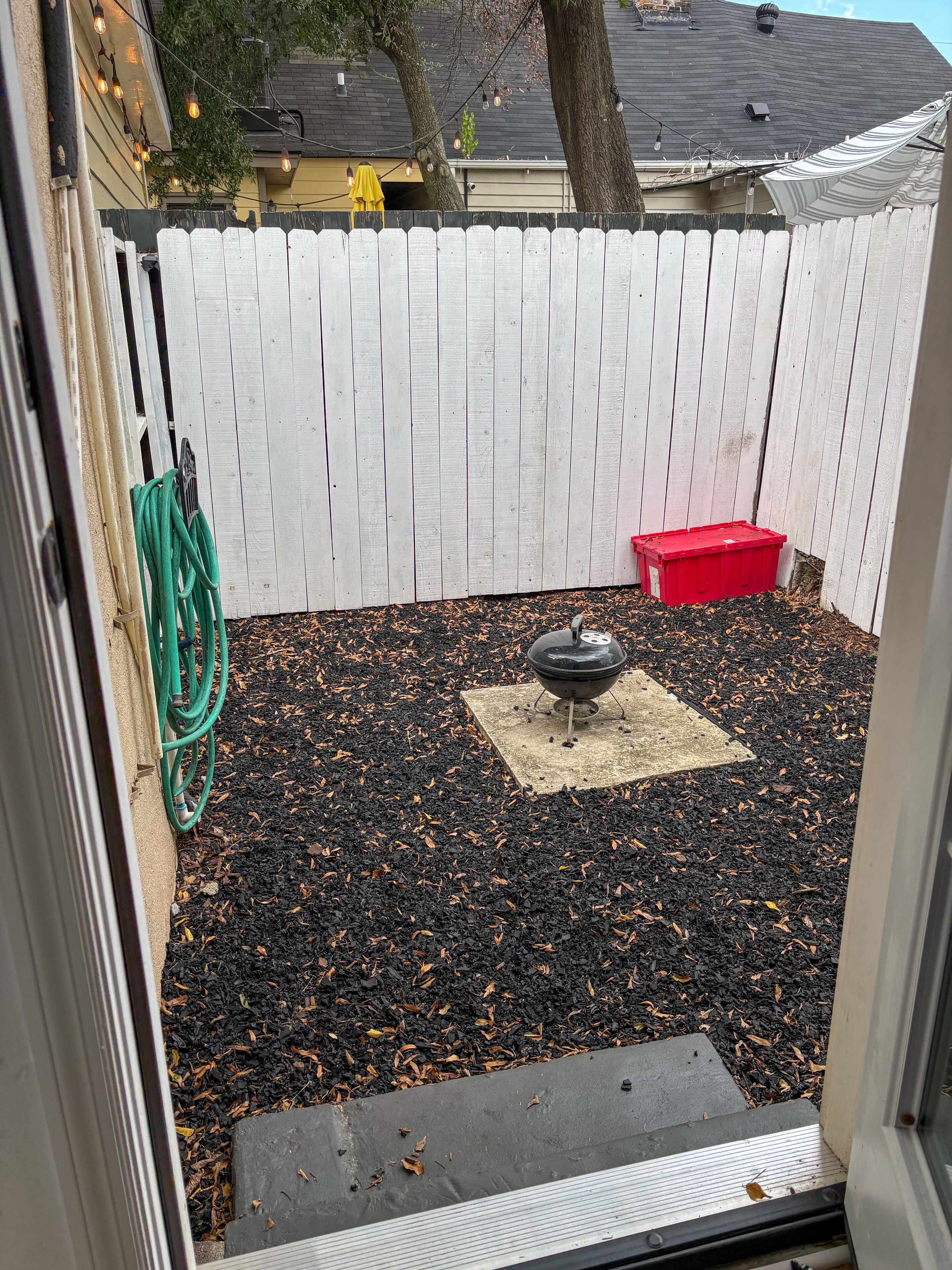 A backyard with a white fence, a barbecue grill, a red container, and a garden hose.
