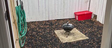 A backyard with a white fence, a barbecue grill, a red container, and a garden hose.
