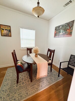 A dining room with a round table, chairs, a vase with dried flowers, a window with blinds, a painting on the wall, and a patterned rug.