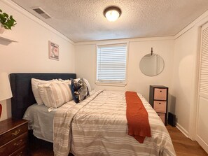 A bedroom with a bed, a dresser, a bedside table, a mirror, and a window with blinds.