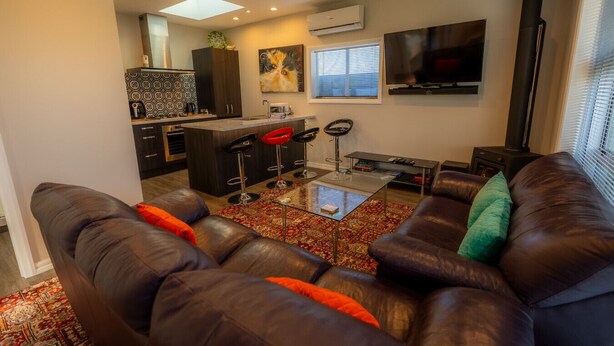 A modern living room with a brown leather sofa, a glass coffee table, and a kitchen area with a bar stool and a painting on the wall. - Cozy Nook Twizel – A Charming Hideaway for Two or a Few (Twizel)