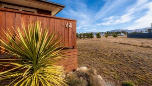 A wooden house with the number 64, surrounded by dry grass and a few trees, with mountains in the background.