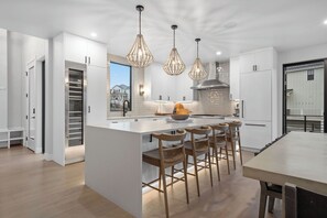 Een moderne keuken met witte kasten, een centraal eiland met barstoelen, hangende verlichting en uitzicht op een huis door het raam.