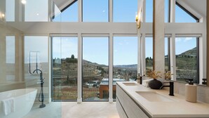 A modern bathroom with a freestanding bathtub, a large sink, and a view of a hilly landscape through large windows.