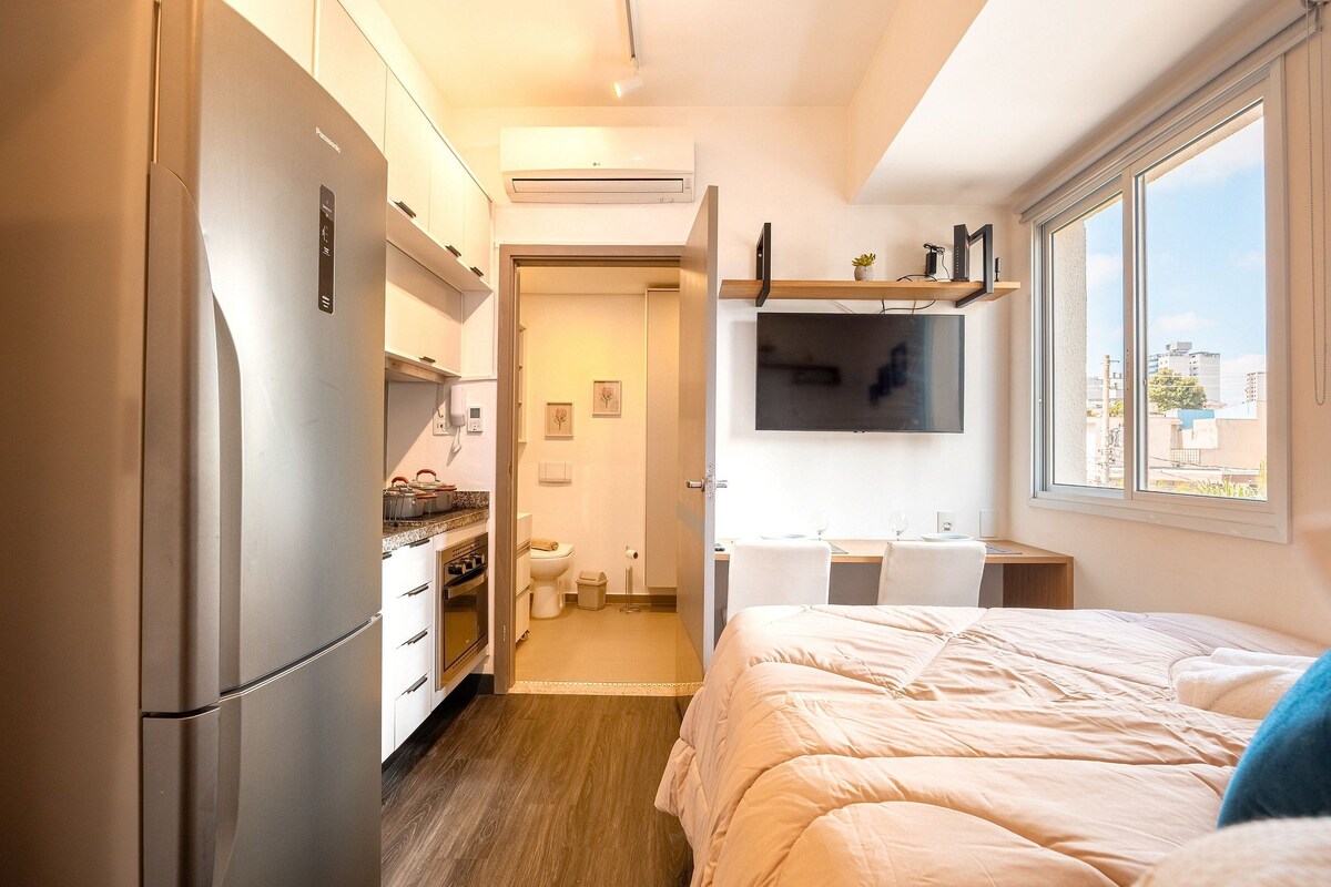 A compact hotel room with a kitchenette, a bed, a TV mounted on the wall, and a window providing a view of the city.