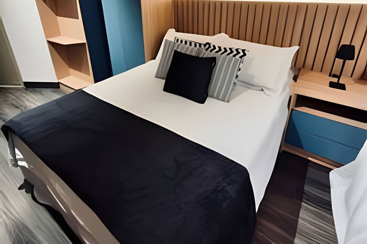 A modern hotel room with a neatly made bed, a bedside table with a lamp, and built-in shelving.
