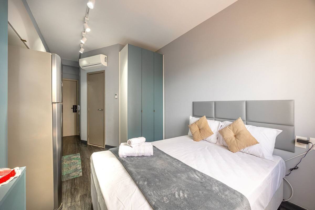 A hotel room with a bed, a grey throw blanket, a white bedside table, and a wardrobe.