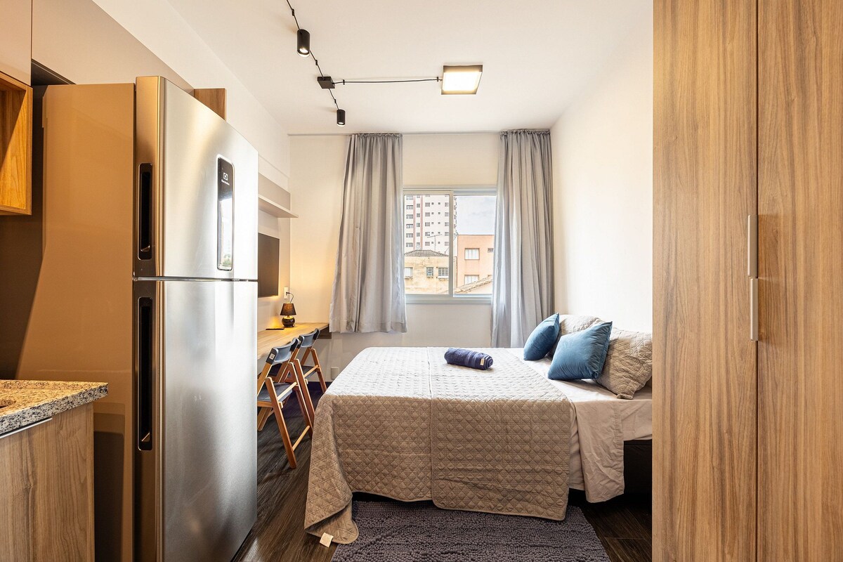 A compact room with a bed, a kitchenette, and a window with curtains.