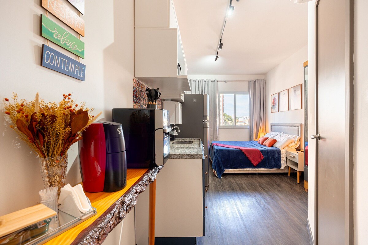Una habitación de hotel moderna con kitchenette, cama con cubrecama azul y rojo, y vista de la ciudad por la ventana.