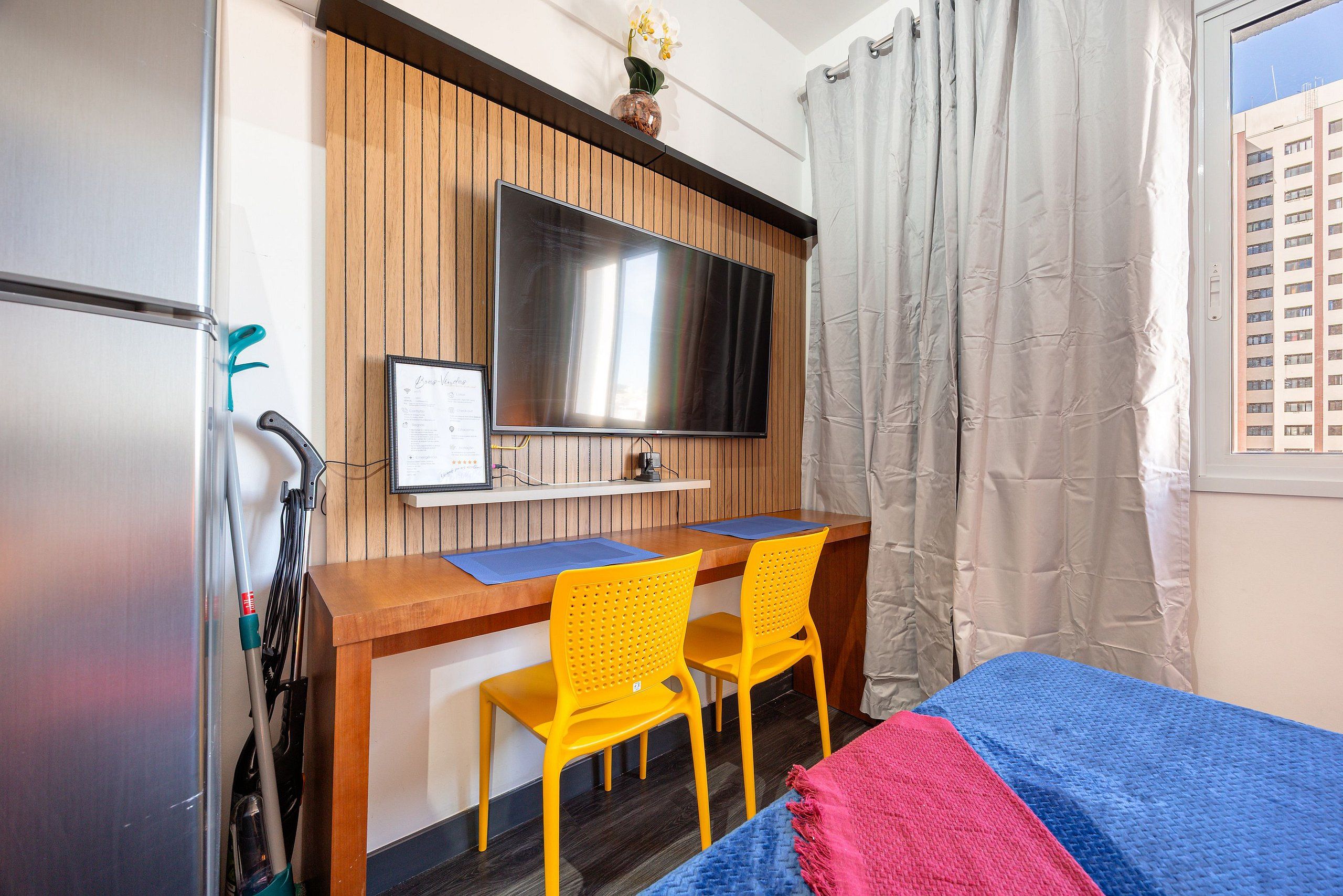Ein Hotelzimmer mit einem Holztisch, zwei gelben Stühlen, einem Fernseher, einem Bett mit blauer und roter Decke sowie einem Fenster mit Vorhängen.