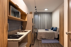 A compact kitchenette with a microwave, sink, and built-in oven. A bed with a quilted cover and pillows is visible in the background. - Estudios Neo Home São Caetano (Sao Caetano do Sul)