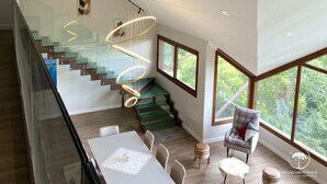 A modern interior with a staircase, dining area, and large windows offering a view of greenery. - Apt Luxo 5 suítes em frente rua torta (Gramado)
