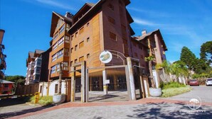A multi-story residential building with a gated entrance, large windows, and a visible sign indicating a location by temporary arrangement. - Apt Luxo 5 suítes em frente rua torta (Gramado)