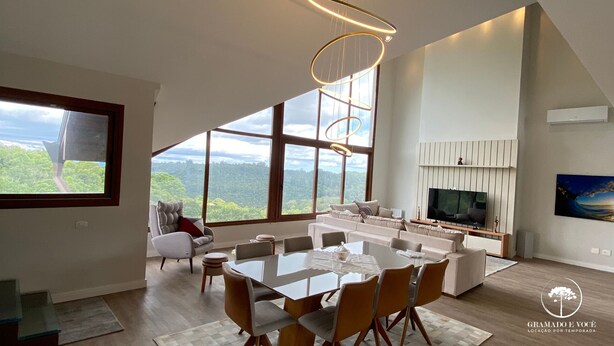 A spacious living area with a large dining table, comfortable seating, a TV, and a scenic view through large windows. - Apt Luxo 5 suítes em frente rua torta (Gramado)