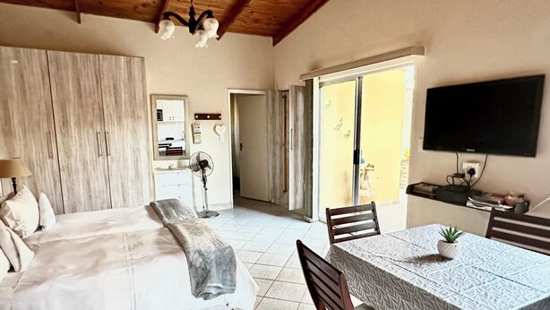 A bedroom with a bed, bedside table, and a fan. There is a dining area with a table and chairs. - Cosy Coastal Escape - Quiet Hideaway 7 minutes from the Sea (Swakopmund)