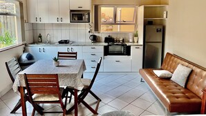 A compact kitchen with white cabinets, a microwave, a stove, and a refrigerator. There is a dining table with chairs and a brown leather sofa. - Cosy Coastal Escape - Quiet Hideaway 7 minutes from the Sea (Swakopmund)