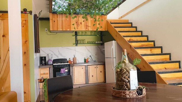 A modern kitchen with a wooden staircase, a dining table with a bottle of wine, and a green wall with hanging plants. - Bottle House (San Felipe)