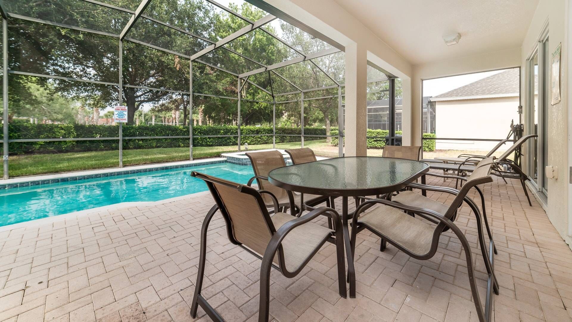 A covered outdoor area with a pool, a round table with chairs, and a view of the surrounding greenery.