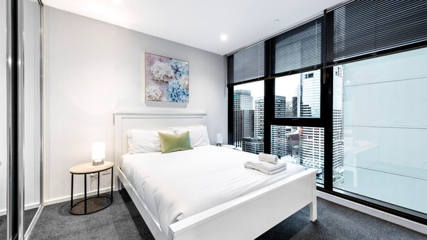 A modern hotel room with a large bed, a bedside table with a lamp, a city view through floor-to-ceiling windows, and a painting on the wall. - Green & Serene - Lush 1BR w Pool & Gym (Melbourne)