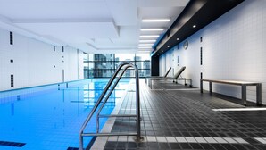 A modern indoor swimming pool with clear blue water, a metal ladder, and lounge chairs along the wall. - Green & Serene - Lush 1BR w Pool & Gym (Melbourne)