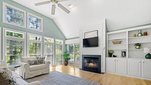 Un salon spacieux doté d’une cheminée, de grandes fenêtres et d’un ventilateur de plafond.