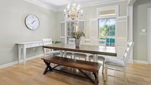 Une salle à manger avec une table en bois, des chaises blanches, un banc, un lustre, une horloge et une fenêtre avec des volets.