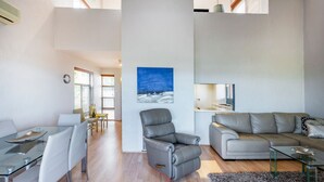 A modern living room with a grey sofa, a glass coffee table, and a leather armchair. There is a painting on the wall and a kitchen area visible through a window. - Como Mezzanine Retreat (Como)