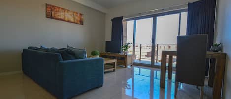 A living room with a sofa, a TV, a small table, and a balcony with a view.