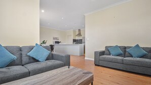 A modern living room with two grey sofas, each with a blue pillow, a wooden coffee table, and a kitchen in the background. - Marvellous McKay 3x2 Close to CBD (Port Hedland)