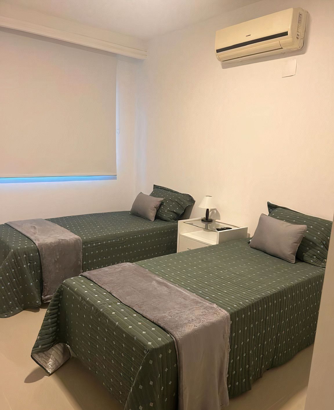 A small, clean room with two beds, a nightstand, a lamp, and an air conditioning unit.