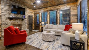 Una sala de estar acogedora con chimenea de piedra, sofás rojo y beige, un otoman redondos y un televisor de pantalla plana montado en una pared de piedra.