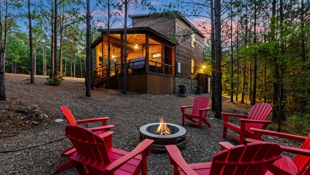 Una cabaña con una fogata y sillas Adirondack rojas en un entorno boscoso.