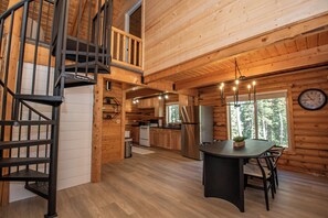 A wooden cabin interior with a spiral staircase, a dining area with a table and chairs, a kitchen with modern appliances, and a large window offering a view of the forest. - Cozy 2-bedroom cabin in tranquil Brian Head (Brian Head)