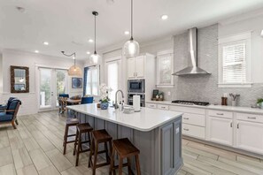 Uma cozinha moderna com armários brancos, uma ilha central com banquetas, iluminação pendente e uma coifa de aço inoxidável.