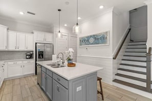 A modern kitchen with white cabinets, a central island, stainless steel appliances, pendant lighting, and a staircase to the right. - Coastal Hip-Nautic by Stay on 30a (Inlet Beach)