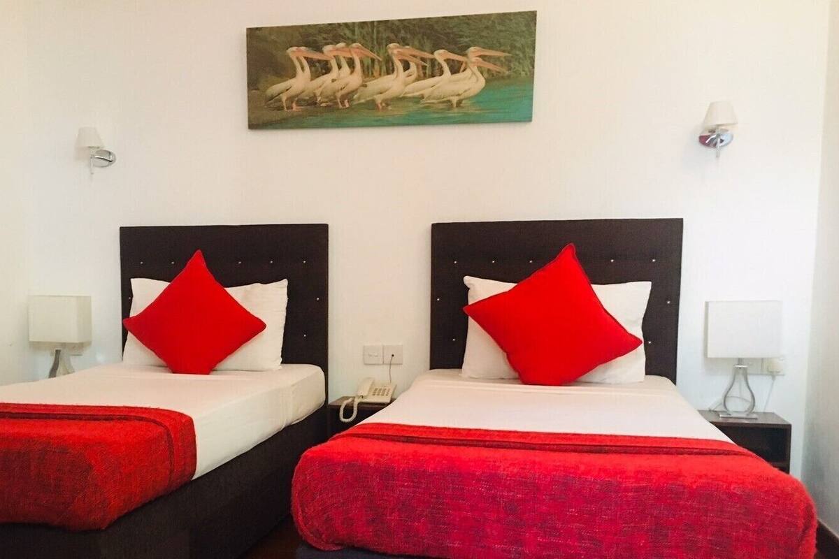 A hotel room with two beds, each with a red blanket and pillow, a painting of birds on the wall, and bedside lamps.