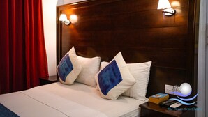 A hotel room with a wooden headboard, two bedside lamps, a bed with white bedding and blue pillows, a nightstand with a remote control, and a red curtain. - The Ocean Pearl Hotel Negombo (Kochchikade)