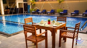 Un área de comedor junto a la piscina con sillas y una mesa de madera dispuestas para dos personas, con vista a una piscina con hidromasaje.