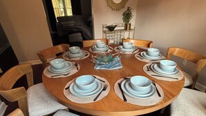 A round wooden dining table with six chairs, each with a light grey cushion. The table is set with blue plates, white napkins, and silverware. - Galactic Oasis Theme Home,  Marvelous 5-bedroom house with AC in Aurora, TV’s (Aurora)
