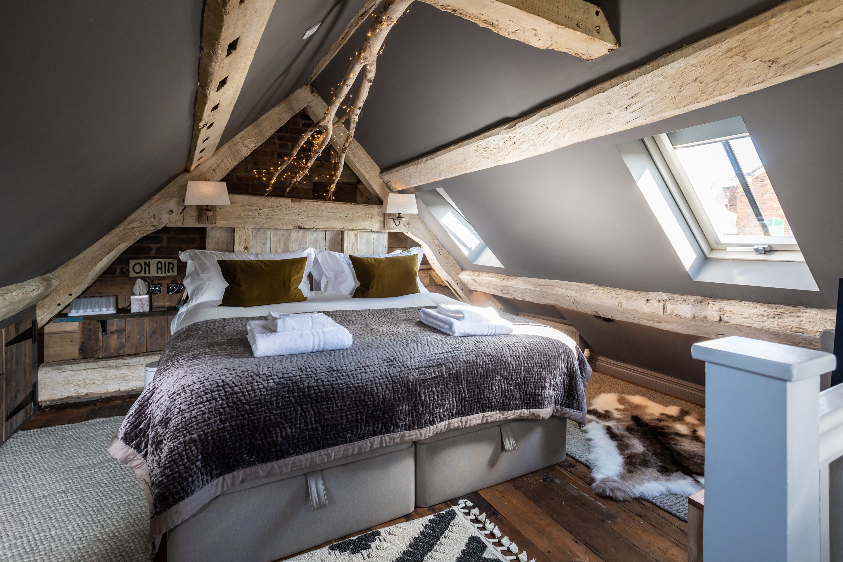 Un dormitorio tipo loft con techo inclinado, vigas de madera a la vista, una cama grande con almohadas, una ventana y una alfombra de piel.